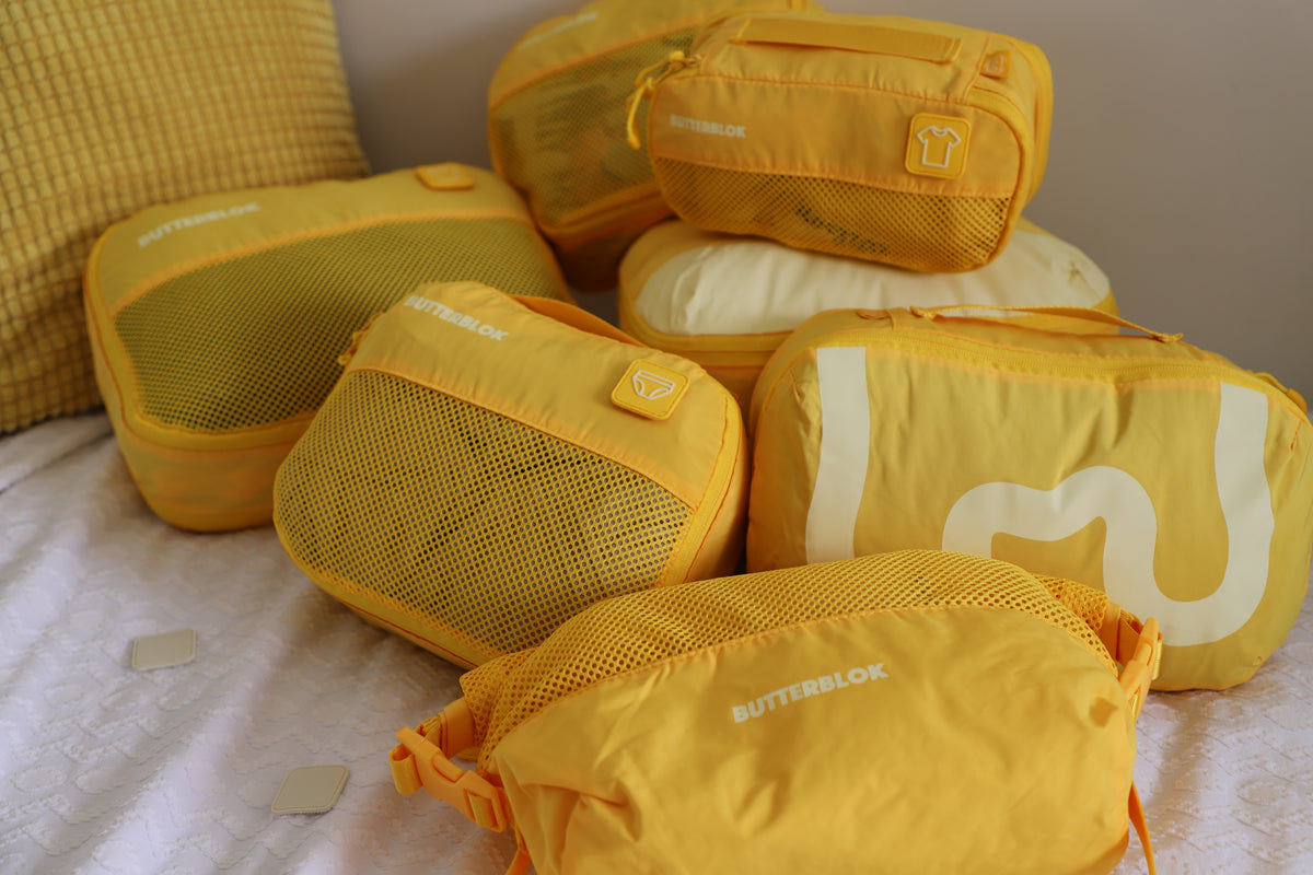 A traveller pulling a suitcase through a whitewashed street, a yellow Butterblok cube strapped on top