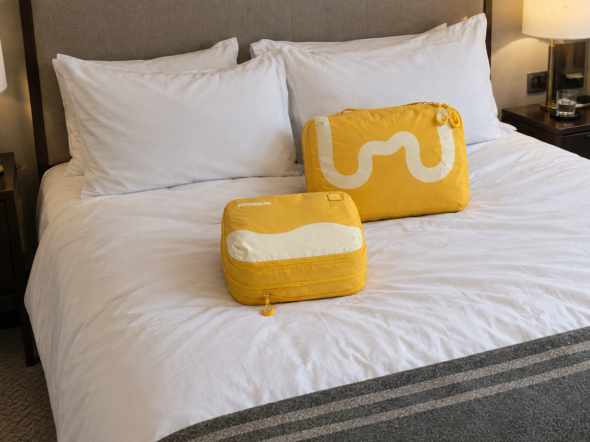 A traveller pulling a suitcase through a whitewashed street, a yellow Butterblok cube strapped on top